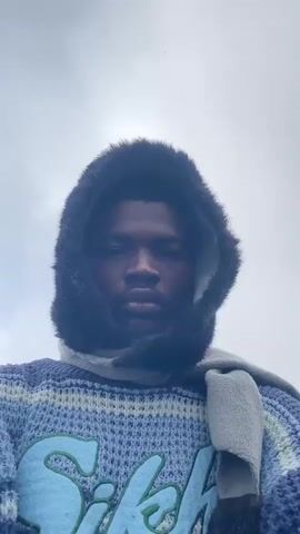 A person is filmed from the chest up. They are wearing a blue and white striped sweater and a gray scarf wrapped around their neck. A black hooded jacket with faux fur lining is pulled up over their head. The person has dark skin and short dark hair. Their eyes are brown. The background appears to be outdoors, with cloudy skies and foliage visible. The person looks directly at the camera with a neutral expression. In some shots, the person's gaze shifts downwards.