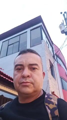 A close-up selfie video shows a man with short, dark hair wearing a black shirt and a camouflage backpack. The background features a two-story building with white siding, large windows, and a red roof. Power lines and a utility pole are also visible against a light sky. The man maintains a neutral expression throughout the video, looking directly at the camera.