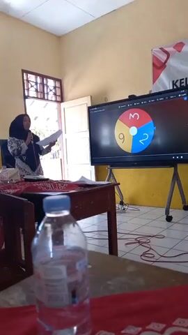 A woman wearing a hijab and a patterned blouse stands in front of a large interactive screen, holding papers and a microphone. The screen displays a spinning wheel divided into three colored sections: red with the number 3, yellow with the number 9, and blue with the number 2. The woman appears to be presenting or teaching, as she looks towards the screen and speaks into the microphone. In the foreground, a wooden table is covered with a red cloth, and a partially filled plastic water bottle rests on it. Another desk is visible in the middle ground, also with papers on it. The room appears to be a classroom or meeting space, with a yellow wall in the background and windows visible on the left. The lighting suggests it is daytime.