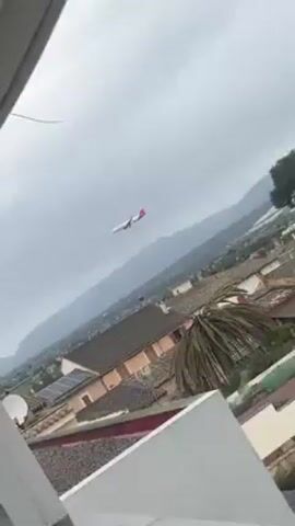 A passenger airplane descends to land on a cloudy day over a cityscape and farmland with mountains in the background. The plane is white with red markings on the tail. The rooftops of buildings are visible below the plane, including a roof with solar panels. The video is shot from an elevated position, possibly a rooftop or balcony. The camera moves to the right, obscuring the plane with the structural support of the building.