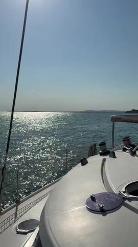 A first-person view from a sailboat shows the bright sun reflecting off the sparkling blue ocean. The edge of the boat’s deck, including a white railing and a dark blue hatch, is visible in the foreground. A black mast extends vertically along the left side of the frame. The horizon is a thin line separating the sea from a light blue sky with a few faint clouds.