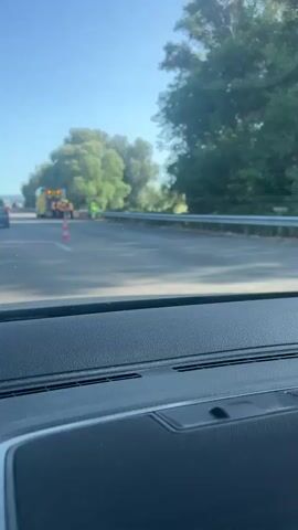 The video shows a highway scene with a fire truck, emergency personnel, and traffic cones. The fire truck is yellow and red, with its back doors open and several individuals in orange vests and helmets working around it. A person in a bright yellow vest stands further down the road, seemingly directing traffic. Orange and white traffic cones are placed on the road. The surrounding environment appears to be a rural area with trees and fields visible in the background. The sky is clear and blue, suggesting a sunny day. The footage is taken from inside a vehicle, looking out through the windshield.