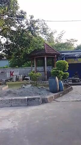 The video begins with a view of a wooden gazebo in an outdoor area with trees and a building in the background. Two large blue barrels are visible next to the gazebo. The camera then pans to the right, showing a strip of shops or stalls. A large red and white banner with text is hanging from the roof of one of the buildings. There are also several smaller signs and advertisements. In the foreground, there is a paved area with a metal barrier and some red stands. As the camera continues to pan, two men are seen interacting at a counter, possibly a gas station or a shop. One man is wearing a white shirt and the other is wearing a dark shirt. There are bottles and containers on the counter. The scene appears to be outdoors, likely in a commercial area.