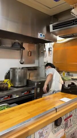 A male chef wearing a black cap, white mask, white shirt, and white apron is working in a commercial kitchen. He reaches up to a stainless steel shelf, retrieves a small blue-patterned bowl, and then uses a long-handled ladle to scoop liquid from a large metal pot on a stove into the bowl. The kitchen features stainless steel surfaces, a ventilation hood, and various kitchen equipment. In the foreground, a wooden counter separates the kitchen area from what appears to be a dining or service area, with patterned wallpaper below the counter.
