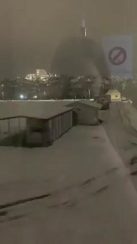 The video shows a view from a window, likely of a moving train or bus, showcasing a snowy landscape at night. A building can be seen covered in snow, along with snowy fields and possibly houses. The lighting is dim, with the glow of city lights visible in the distance. The camera movement suggests that the vehicle is moving, causing the view to change. The presence of a sign with a "no" symbol, possibly a "no smoking" sign, can be observed near the window.