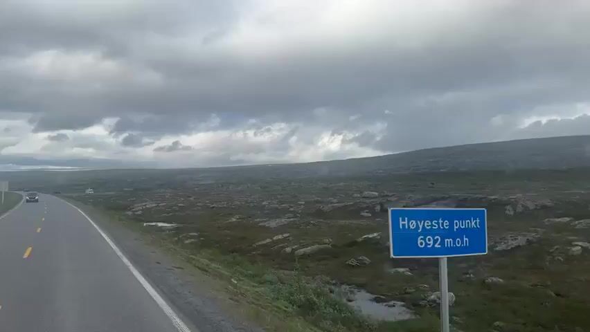 This media file shows a scenic drive through a vast, rocky, and sparsely vegetated landscape under a dramatic, cloudy sky. The foreground features a paved road with yellow lane markers, curving to the right. The road appears to be in good condition. To the right of the road is a gentle slope covered in low-lying green vegetation interspersed with numerous grey and brown rocks of various sizes. The terrain beyond the road is a wide, undulating plateau covered in similar vegetation and rocks, stretching towards a distant, hazy horizon. Small bodies of water, possibly puddles or shallow ponds, are visible in the rocky terrain.

In the distance, along the road, a few vehicles are visible, suggesting this is a route used for travel. These appear to be cars or vans. The sky is dominated by heavy, grey clouds, indicating overcast or potentially stormy weather. There are hints of sunlight breaking through in some areas, creating shafts of light, and the overall lighting suggests it is daytime, likely late afternoon or evening due to the muted light and the dramatic cloud formations.

The landscape evokes a sense of remoteness and rugged beauty, characteristic of mountainous or subarctic regions. The presence of the road and vehicles indicates human activity within this wild setting. There are no visible people in the frame, but the activity is clearly a journey or travel by road. A white signpost with horizontal lines is visible on the left side of the road in the mid-ground. The overall mood is one of atmospheric grandeur, with the natural landscape taking center stage under a dynamic sky.