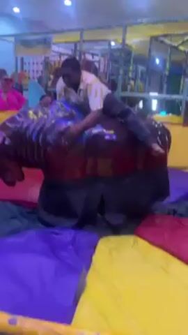 The video shows a young man riding a mechanical bull, which is dark brown with multicolored spots and has long, curving white tusks. The bull is set up on a padded floor made of purple, yellow, and red segments. The man is wearing a patterned yellow and white short-sleeved shirt and dark pants. He struggles to hold on as the bull moves, initially lying across its back but then pulling himself up to sit more upright. A woman in a bright pink outfit can be seen standing to the left of the frame, apparently watching and possibly filming the scene. A young girl is visible behind the woman, also watching. The scene appears to be in an indoor play area with a climbing structure visible in the background.