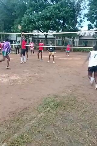 The video shows a volleyball game in progress on a dirt court outdoors. There are about a dozen players, mostly dressed in athletic attire including jerseys, shorts, and sneakers, distributed on either side of a standard volleyball net. Some players are moving, while others are stationary, anticipating the ball. In the background, there are green trees and buildings. The camera angle is slightly shaky.