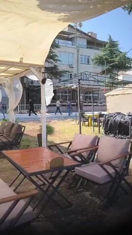 The video displays an outdoor seating area of what appears to be a cafe or restaurant. In the foreground, there are several tables and chairs with cushions, suggesting an established seating arrangement. A wooden table with a metal frame is positioned prominently in the center, with a small object on it. Further back, more seating arrangements are visible, including lounge chairs and smaller tables. The area is partially covered by a light-colored canopy. In the background, a few individuals are walking around. One person in a dark jacket is seen walking across a grassy area. Two other individuals are standing and facing each other near a structure. Behind them, a building with multiple windows and balconies is visible, along with trees and streetlights. The overall impression is of an open-air dining or relaxation space.