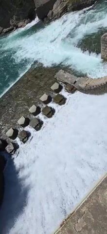 The camera pans up to reveal a view of a river, flowing from the mountains on the horizon, moving across a rocky landscape, and cascading over a small, man-made dam.  The dam is made of concrete, with some large, geometric concrete blocks at the base.  The water roars as it flows over the dam, creating a white, foamy texture. Trees and vegetation line the banks of the river. The sky is blue with some light cloud cover.
