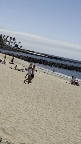 A video shows a sandy beach with people sunbathing and walking. In the background, the ocean is visible with a breakwater and a distant lighthouse. The sky is clear with some clouds. Several individuals are lying on towels on the sand, while others are walking along the shoreline. Some people are seen interacting with each other, and there are children playing near the water. The overall scene depicts a typical sunny day at a beach resort.