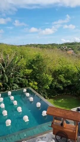 The video shows an outdoor setting in a tropical location. A pool with floating white lanterns is visible, along with a wooden cart and a lounge area with blue cushions. Beyond the pool and lounge area, there is lush green vegetation. In the distance, rooftops of buildings can be seen, along with the ocean. The camera angle changes to show the scene from under an open glass window in a wooden structure. The ocean and cliffside are clearly visible. The weather is sunny with a blue sky and scattered clouds.