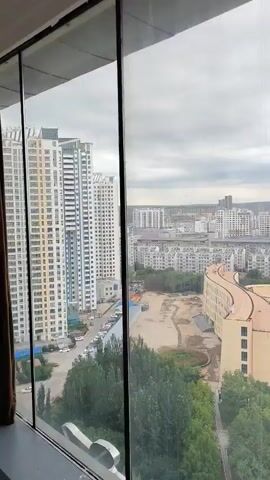 The video shows a panoramic view of a city from a high vantage point, possibly a window or balcony. The scene transitions from a closer view of buildings and trees to a wider perspective of a city street. In the foreground, there are tall apartment buildings and greenery. As the view pans out, a wide road with multiple lanes is visible, lined with trees and buildings. Traffic, including cars and buses, is moving along the road. The sky is overcast. The video appears to be a time-lapse or a slow pan, showing the city in daylight. There are no discernible people interacting in the video; the focus is on the urban landscape.