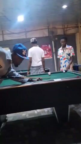Here's a description of the video:

The video shows a pool hall setting with three visible pool tables. Three men are present: one in the foreground wearing a grey jacket and a blue baseball cap, another in a white T-shirt and patterned shorts standing by the second pool table, and a third man wearing a brightly patterned shirt near the back.

The man in the blue cap is taking a shot at the pool table closest to the camera. The pool hall has overhead lighting, a bare concrete floor, and posters on the walls. Another man in a yellow shirt is seen walking behind the first two tables.