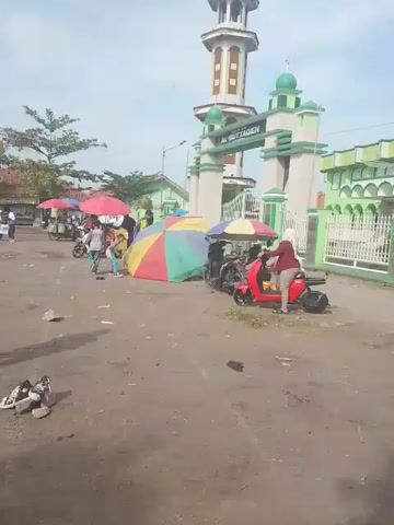 The video shows a street scene near the Masjid Jami Al Muttaqien mosque in an unidentified location. Several motorcycles and people are visible. Children in what appear to be school uniforms are near food vendors' carts. Some adults are interacting with children, and others are walking around. The mosque has a distinctive green and white color scheme. The ground appears to be dirt or gravel. The sky is partly cloudy.