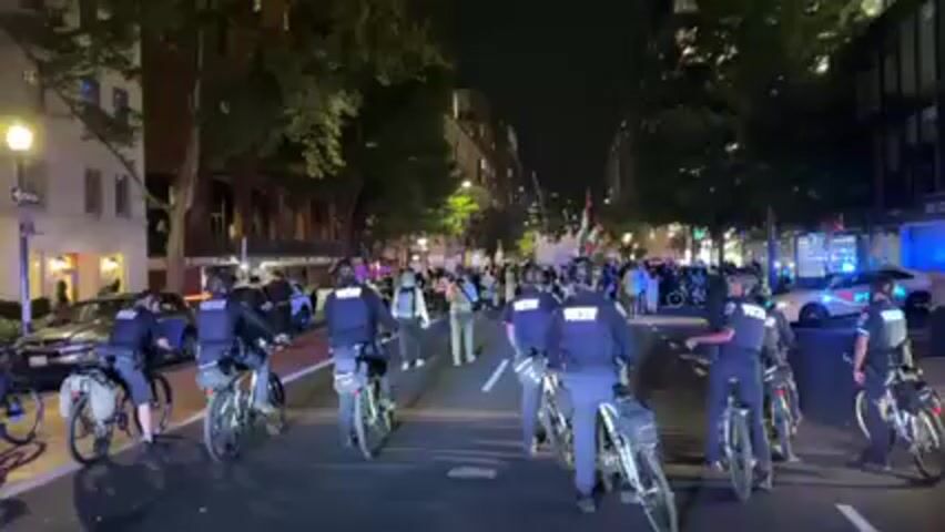 A nighttime street scene shows a line of police officers on bicycles facing a crowd of people. The officers are wearing dark uniforms with "POLICE" printed on the back. Some are also wearing helmets. They are positioned in the middle of the street, forming a barrier between the camera and the crowd. In the background, the crowd is diverse and appears to be protesting, with some individuals holding flags. A police car with flashing lights is parked on the right side of the street, behind the officers. The street is lined with buildings and trees, and streetlights provide illumination. The overall atmosphere is tense due to the presence of both police and protesters. A loud noise, possibly a firework, occurs around 0:01.