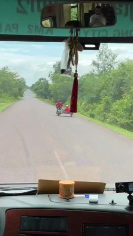 A motorcycle with a sidecar travels on a paved road. The motorcycle is ridden by a person wearing a red hooded jacket. A second person, also wearing a red jacket and a white cap, sits in the sidecar. The motorcycle is pulling the sidecar, which appears to be made of metal with a light blue panel on the side. The road is surrounded by lush green trees and vegetation. The sky is cloudy. The camera appears to be mounted inside a vehicle, looking out through the windshield. Various objects are visible on the dashboard of the vehicle, including a small camera, a container, and what looks like a car stereo. A decorative tassel hangs from the rearview mirror.