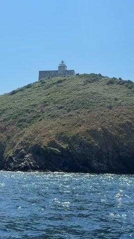 The video shows a lighthouse on a grassy hill overlooking the sea. The sky is clear and blue. The water in the foreground is choppy and reflects the sunlight. A few birds can be seen flying in the distance. The lighthouse is a white, rectangular building with a smaller tower on top. The hill is covered in green vegetation and rocky outcrops.