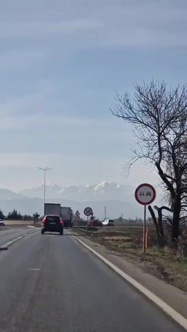 The video shows a road with several vehicles, including a dark-colored SUV, a red car, and two large semi-trucks with trailers. The SUV is the closest to the camera, driving on the right side of the road. Further ahead, there's a truck with the word "LANDWEHR" visible on the side of the trailer, and another truck behind it. Several road signs are visible, including a "30" speed limit sign, a roundabout sign, and a "50" speed limit sign. On the right side of the road, there's a round sign with a red border and a white center, seemingly indicating a prohibition or warning. In the background, a mountain range with snow-covered peaks is visible under a sky with some clouds. The road appears to be passing through a relatively flat, rural area. The video is taken from a moving vehicle.