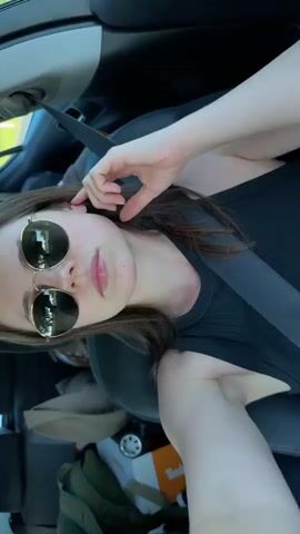 A woman with shoulder-length brown hair and fair skin sits in the driver's seat of a car. She is wearing round sunglasses with gold frames, a black ribbed tank top, and a seatbelt. She has pink lips. The car's interior is mostly black and white. A Timberland shoebox is visible on the passenger seat. Bright sunlight casts shadows across her face and the car's interior. The camera angle is slightly tilted. The video is short and static.