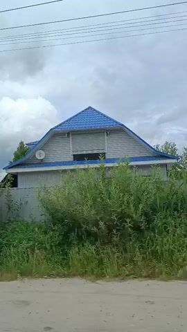 The video captures a drive through a rural area, likely in Снопок Новый, Russia, on a cloudy day. The scene alternates between exterior shots of a road with trees and power lines, and interior shots from the perspective of a car's dashboard.

The exterior shots show a dirt or gravel road alongside a paved road. Power lines run overhead, supported by wooden poles, one of which has electrical equipment attached. Dense green foliage lines the roads, with trees and bushes forming a verdant backdrop. A white SUV is visible on the left side of the road in one shot. In another, a small transformer station with an air conditioner unit is visible behind a metal fence. A light blue house with a blue roof is partially visible on the right.

The interior shots focus on the car's dashboard, adorned with a vibrant bunch of purple lupine flowers. Through the windshield, the road stretches ahead, with more trees and power lines on either side. Other vehicles can be seen on the road in the distance. The sky is overcast with dramatic, dark clouds, suggesting the possibility of rain or a storm. A faint contrail is visible in the sky in one of the interior shots.

The overall mood is serene and contemplative, enhanced by the natural beauty of the landscape and the simple adornment of the car's interior. There is no discernible human activity beyond the act of driving. The time of day appears to be daytime, given the ambient light, despite the cloudy weather. No text is visible in the video.