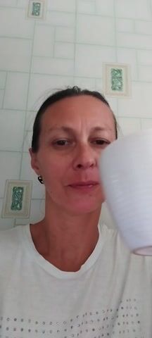 The video shows a person with dark hair, wearing a white t-shirt, against a tiled background. The person is holding a white mug and drinks from it. They then lower the mug and stick out their tongue. The video is captured in portrait orientation.