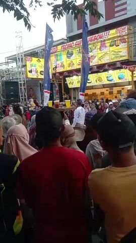 A crowd of people stands outside a building with signs for "SONIC CHICKEN" and a bank. Some people are wearing headscarves. There is a stage with speakers and a ladder to the left. People are looking towards the building and some are holding up phones. It appears to be an outdoor event or gathering.