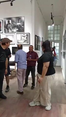 The video shows a group of people standing and chatting in what appears to be an art gallery. The gallery has white walls and is lined with framed photographs.  Several people are in small groups, talking and looking at the artwork.  One woman is holding a champagne glass, and a man holds a drink can. The floor is wooden and has a reddish-brown tone.  Light fixtures are visible on the ceiling.  Some people are walking along the hall.