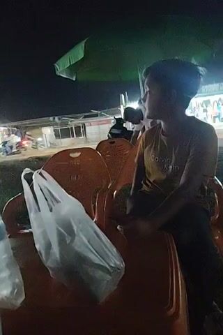 A young boy is seated on an orange plastic chair outdoors at night. He is wearing a brown t-shirt with the words "Calvin Klein" on it. To his left is a white plastic bag resting on another orange chair. There are several other chairs around him. Above him is a green umbrella providing some cover. In the background, there is a truck driving by with its lights on. A few other people are visible in the background, including one person in a white shirt. The location appears to be a roadside or an outdoor eating area. The boy seems to be talking or observing the surroundings.