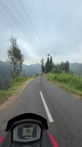 This is a first-person view video taken from a motorcycle as it travels along a paved road in a mountainous, rural area. The road curves to the left, and on either side, there is lush green vegetation and tall trees. In the distance, rolling hills and more trees are visible under a cloudy sky. There are power lines running overhead. The motorcycle's digital display shows a speed of 39, then 40, and finally 41. Another motorcycle with a rider is visible ahead on the road. Yellow and black striped markers are placed along the side of the road. The overall atmosphere is serene and natural.