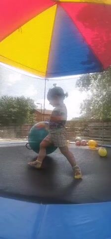 A child is jumping on a trampoline with several balls. The trampoline has a colorful canopy overhead. The child is wearing a patterned shirt and shorts. The child is interacting with a large green ball. Other balls of various colors are scattered on the trampoline. The background shows trees and a fence.