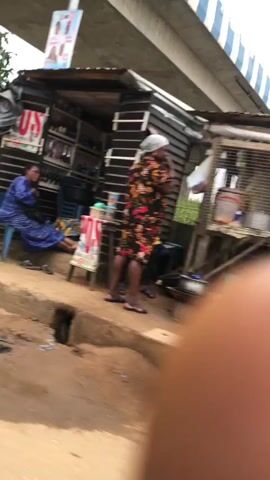 The video shows an outdoor scene under an elevated concrete structure. In the foreground, a blurred finger is visible. To the left, a makeshift wooden and corrugated metal stall is present with a person inside. Further right, another stall is seen with a person in a blue shirt seated. Campaign posters with images of people are affixed to a pillar. A yellow vehicle component is also visible on the left side of the frame. The ground appears to be dirt and gravel.