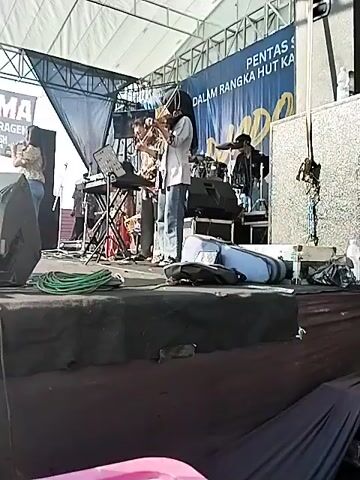 A live band performs on an outdoor stage under a white tent. The stage is elevated with dark fabric covering the sides. The band consists of a female vocalist wearing jeans and a patterned shirt, a keyboardist with a music stand, a violinist wearing a hijab and jeans, and a drummer partially visible in the background. Speakers, cables, and instrument cases are scattered on the stage. A banner in the background reads "PENTAS SENI DALAM RANGKA HUT KA." A sign on the left of the stage displays "PANAMA PUTRA SRAGEN".