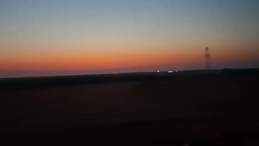 The video shows a wide outdoor landscape at dusk or dawn. The sky transitions from a pale blue at the top to a vibrant orange and red near the horizon, indicating the sun is low. A dark, blurry line of trees or hills separates the sky from the foreground. In the distance, a tall, thin transmission tower with lights is visible. The foreground is very dark and blurry, suggesting it's either nighttime or the viewer is looking from inside a moving vehicle at night. There are some faint lights on the right side of the frame which may be from a vehicle's dashboard or a building. The overall impression is of a vast, open space under a colorful twilight sky, possibly viewed from a moving car.