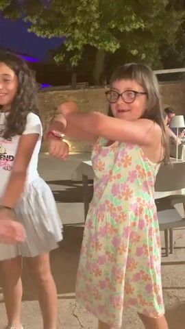 Two young girls are dancing outdoors at night. The girl on the left has dark curly hair and wears a white t-shirt with a graphic and a silver pleated skirt. The girl on the right has brown hair, wears glasses, and is dressed in a light-colored floral print dress. They are both moving their arms in unison, appearing to follow a choreographed dance. The background shows outdoor seating areas with tables and chairs, illuminated by ambient lighting and some spotlights on a stone wall. Trees are visible above. The overall atmosphere appears to be a casual gathering or party.