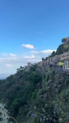 The video shows a scenic view of a Mediterranean landscape on a bright, sunny day. The camera pans across a hillside village with traditional stone houses clustered on the slope. Lush green vegetation covers the hill, and the blue sea extends to the horizon, merging with a clear sky dotted with fluffy white clouds. The scene captures the serene beauty of a coastal Mediterranean setting. The camera angle is slightly low, looking up at the village and the vast seascape beyond. There is faint background conversation.