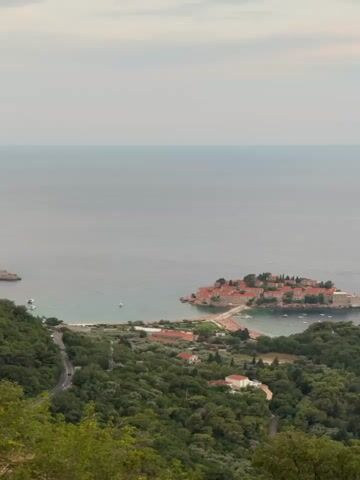 This video shows a high-angle drone shot of a coastal village with an island connected by a causeway. The island has red-tiled roofs and stone buildings, and it is surrounded by the sea. The mainland coastline has lush green hills with scattered houses and a road winding through them. The water is calm, and the sky is overcast. The overall scene suggests a serene and picturesque location.