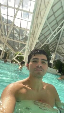 A shirtless man with dark hair stands in a turquoise swimming pool, recording himself. The pool is indoor and is covered by a high, glass, geometric roof. Other swimmers are visible in the background. Green plants are visible along the back edge of the pool.