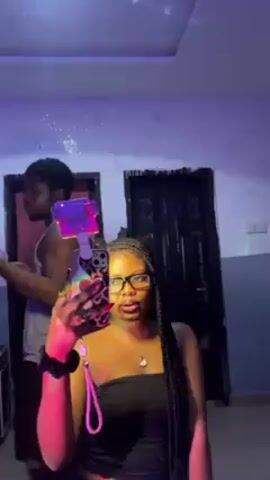 A woman with dark hair in long braids and wearing glasses, a black strapless top, and a necklace is holding a smartphone with a pink phone grip. She is standing in front of a mirror and recording a video. In the background is a man wearing a tank top, standing near a door. The room has a white wall, a dark-colored door, and a ceiling fixture that projects light onto the ceiling. There are flashing disco lights illuminating the scene in pink, green and blue hues.