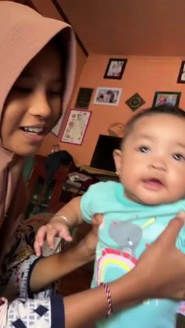 A person wearing a hijab is holding a baby in a teal onesie with a rainbow and elephant graphic. The baby is looking towards the camera with wide eyes. The room appears to be a living room, with framed pictures on the orange wall and a fan in the background. The person holding the baby is wearing a patterned shirt. The video is shaky, suggesting it was recorded handheld.