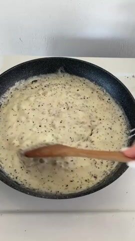 The video shows two close-up shots of cooking. The first shot, from approximately 00:00 to 00:01, shows a black non-stick frying pan with a creamy sauce being stirred with a wooden spoon. The sauce appears to contain finely chopped red onions and black pepper. Bubbles are visible in the sauce, indicating it is hot and simmering. The second shot, from approximately 00:01 to 00:02, shows a stainless steel pot with a glass lid. Inside the pot, spiral-shaped pasta is being cooked in water. Steam is visible condensing on the inside of the glass lid. Both cooking vessels are placed on a white stovetop, with visible marks and residue on the stovetop surface.