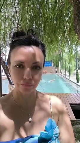 A woman is taking a selfie in front of a swimming pool. She is wearing a blue and white patterned scarf and a necklace with a pearl. She has dark hair pulled up in a bun and is wearing earrings. Behind her, there is a long, narrow swimming pool with blue water and a wooden deck. There are trees with green leaves providing shade over the scene. In the background, there are people walking near the pool. The overall environment appears to be an outdoor recreational area.