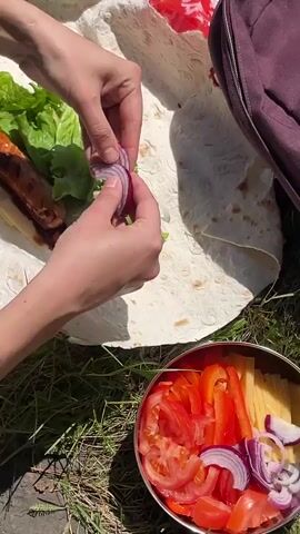 A person's hands are shown assembling a meal on a large piece of flatbread, likely lavash, in an outdoor setting in Sliznevo, Russia. The flatbread already contains a grilled sausage and slices of cheese. The hands are carefully adding fresh green lettuce and thinly sliced red onion to the ingredients.

To the lower right of the frame, a metal bowl holds an assortment of chopped fresh vegetables, including red tomatoes, red bell pepper strips, and more red onion, from which the person takes additional onion to add to the wrap. The ground beneath the food preparation is covered in vibrant green grass, indicating a natural, outdoor environment, possibly for a picnic or barbecue. The bright daylight is evident from the strong shadows cast on the scene. In the background, a red plastic bag with partially visible text, appearing to be "Coba," is seen next to a purple bag or backpack. The video captures the focused and careful preparation of a fresh, portable meal outdoors.
