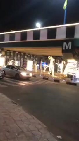 The video depicts a street scene at night, with a footbridge illuminated with string lights spanning the road. Several cars are visible driving on the road. Decorative light displays, including figures resembling reindeer, are set up alongside the road beneath the bridge. Pedestrians are walking in the vicinity of the light displays. The footbridge has a checkered design on top and a prominent letter "M" on its side. A Christmas tree lit with blue lights is seen in the distance through the footbridge. A flag flutters from the bridge, adding to the visual complexity.