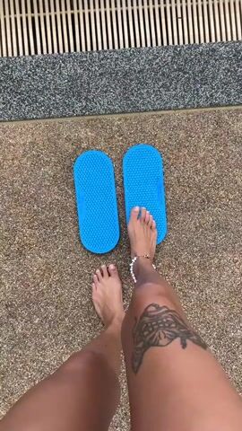 The video shows a person's legs and feet from a top-down perspective. The person is standing next to a pool, with a grate visible in the upper part of the frame. The person is wearing two blue flip-flops with raised bumps, and one foot is already placed on one of the flip-flops. The person then places their other foot onto the second flip-flop. The person has a tattoo of a butterfly on their right thigh and a beaded anklet on their left ankle. The ground around the pool is a rough, speckled surface.