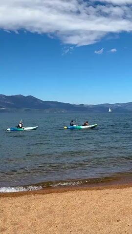 The video shows a wide shot of a calm body of water with a sandy beach in the foreground. Two kayaks are visible on the water. The kayak on the left is white and occupied by one person wearing a white hat and a light-colored shirt. The kayak on the right is colorful (blue, green, and yellow) and occupied by two people. The person in the front of this kayak is wearing a dark shirt and appears to be paddling, while the person in the back is wearing a light-colored shirt and a hat, possibly relaxed or observing. In the background, mountains line the horizon under a bright blue sky with scattered white clouds. A sailboat is visible in the distance on the right side of the frame. The water is clear and reflects the sky. Gentle waves lap at the sandy shore.