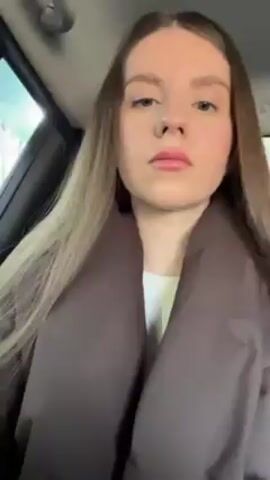 A woman with long brown hair and light complexion is in a car. She is wearing a brown coat and a white top. She opens a small, pink lip gloss container with an orange top. She applies the lip gloss to her lips. The background shows the interior of the car and a glimpse of the outside environment.