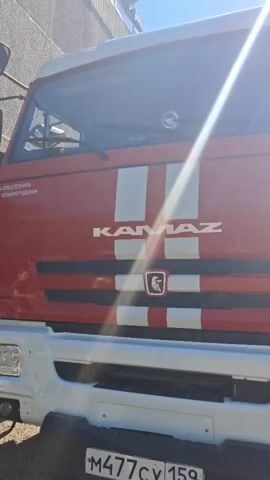 The video shows the front of a red and white Kamaz fire truck parked outdoors on a sunny day. The truck has a large Kamaz logo on the front, as well as some text in Russian that appears to be related to fire services. The driver's side door is open, and several people, including children and adults, are gathered around the truck, some appearing to look inside the cab. A few people are wearing what look like uniforms. The overall scene suggests a public event or an open house related to the fire department.