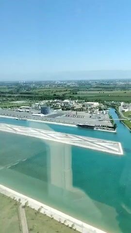 An aerial view captures a man-made waterway with turquoise water, bordered by concrete structures and green grassy areas. Beyond the waterway, a complex of buildings, including a large hotel and other structures, is visible, surrounded by fields and trees. The sky is clear blue, and distant hills can be seen on the horizon. The video pans slowly across the scene, offering a sweeping panorama of the resort-like environment.