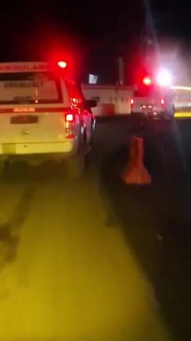 The video shows the rear of a white ambulance with "AMBULANS" and "EMERGENCY" written on it. The ambulance is driving at night, indicated by the dark sky and illuminated lights. There are red and blue flashing lights on the ambulance and other vehicles in the distance. The road appears wet, reflecting the lights. Traffic cones are visible on the right side of the road. The camera perspective is from a vehicle following the ambulance.
